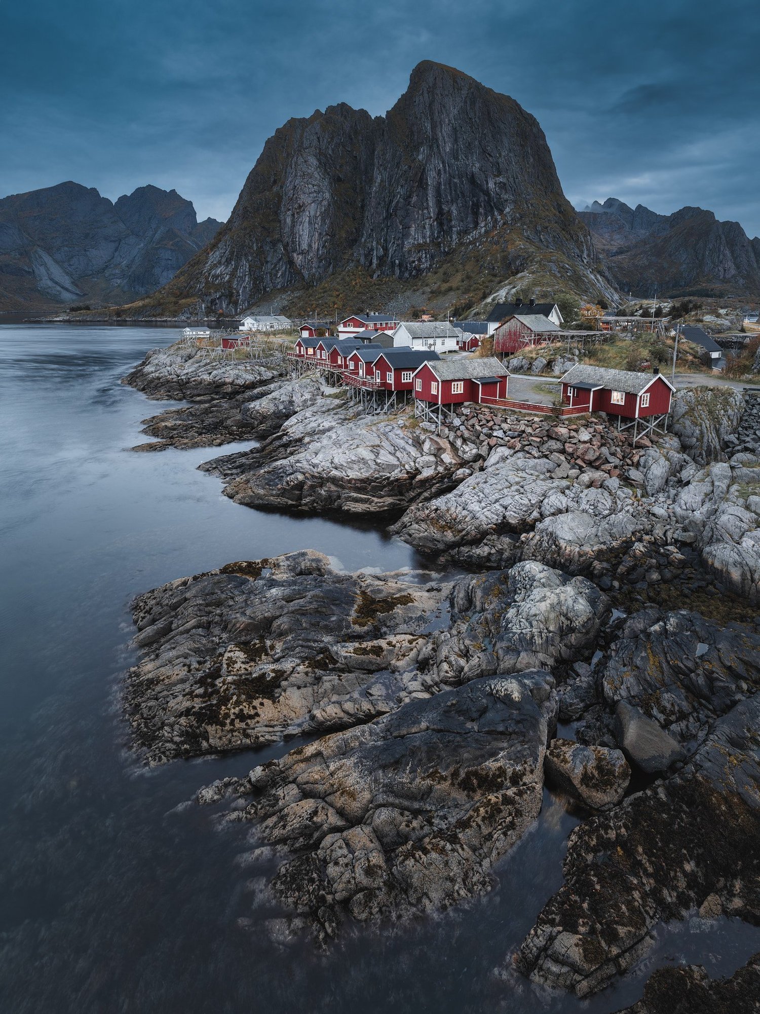 Rote Küstenhäuser auf Stelzen stehen am Wasser vor den Bergen der Lofoten.