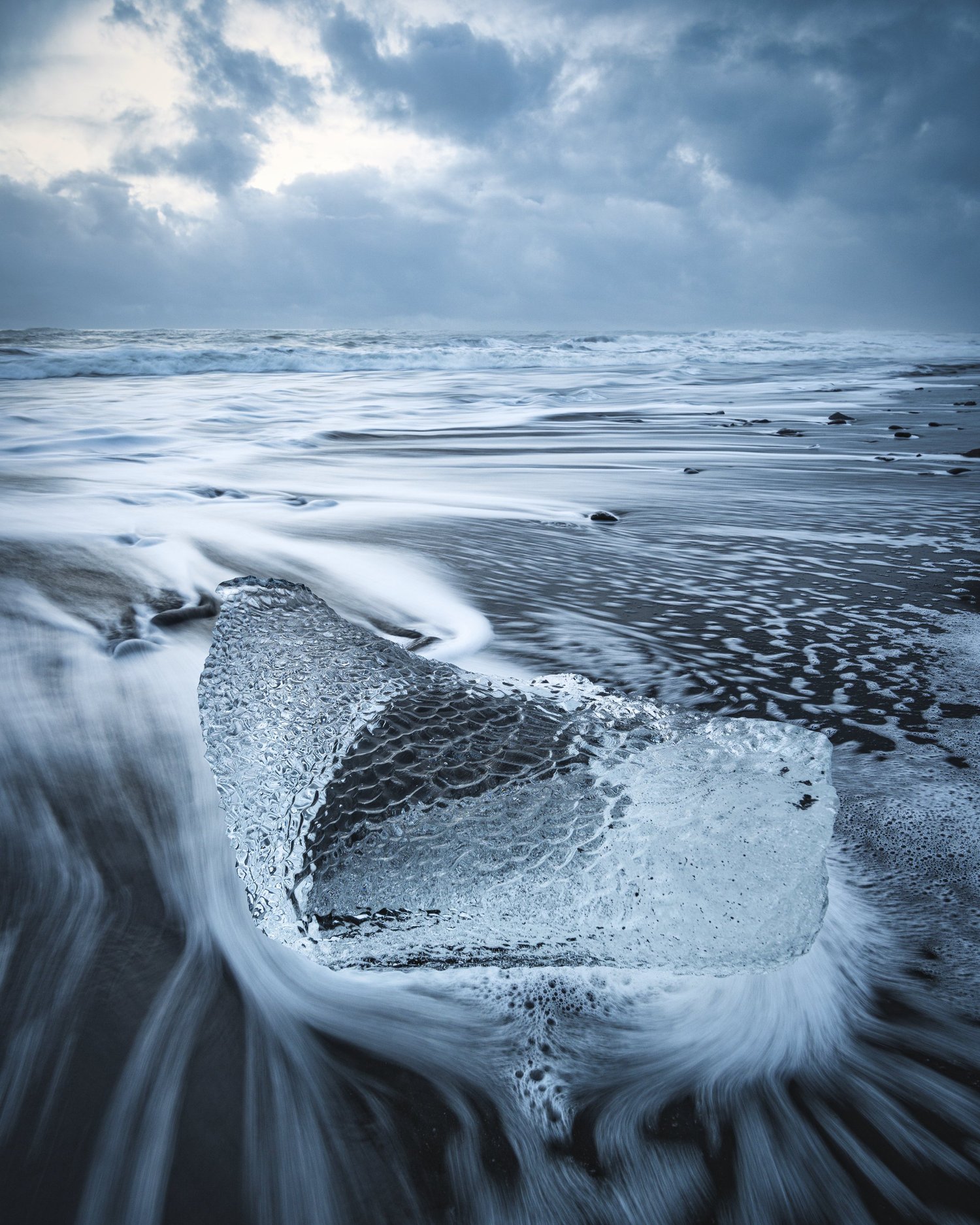 Ein klar geformtes Eisstück liegt am schwarzen Strand, während Wasser an ihm vorbeiströmt.