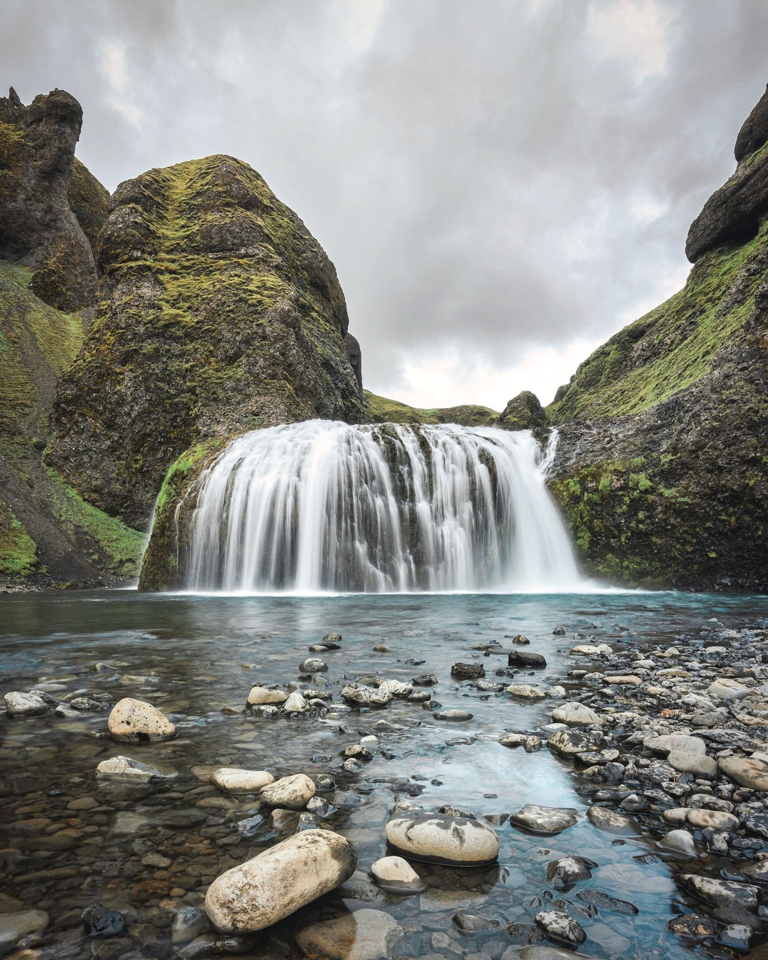 Ein Wasserfall fliesst durch ein grünes Tal über moosbewachsene Felsen und glatte Steine.